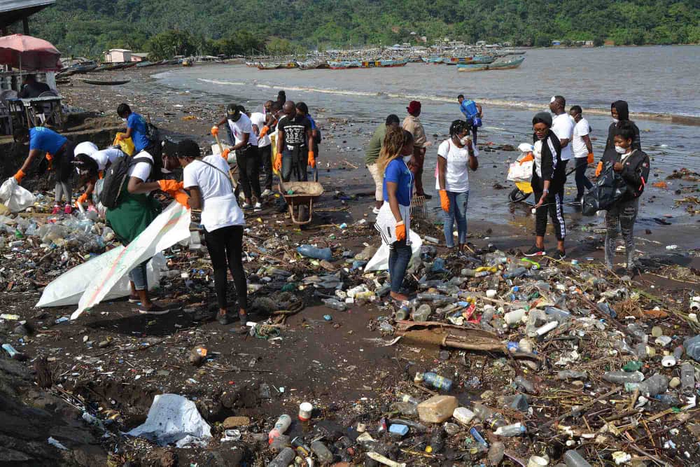 A large group collecting plastic waste from a beach strewn with bottles, with fishing boats offshore.
