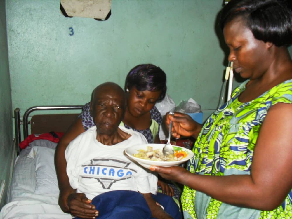 Two women helping an older man with a bowl of food at his bedside.